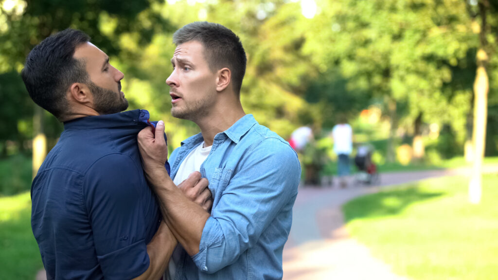 Two men are in a confrontation. The first man is committing assault; he has grabbed the second man by the front of the shirt and is threatening him. The second man looks frightened.